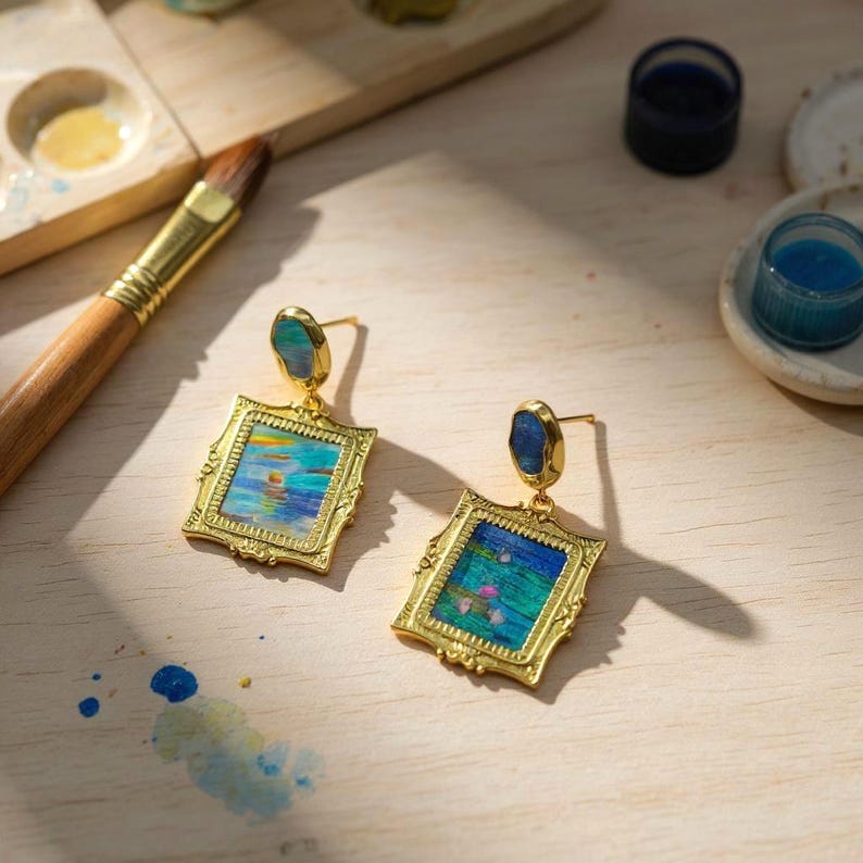 May include: A pair of gold-toned earrings featuring rectangular frames with ornate edges. Each earring displays a miniature impressionist-style painting of water lilies and blue tones, topped with a small, smooth, dark blue oval stone.
