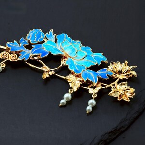 Pode incluir: Um acess&oacute;rio de cabelo em tom dourado com motivos florais e folhas esmaltadas em azul vibrante. A pe&ccedil;a &eacute; acentuada com pequenas p&eacute;rolas artificiais brancas pendentes suspensas em delicados caules dourados, criando um design bot&acirc;nico detalhado.