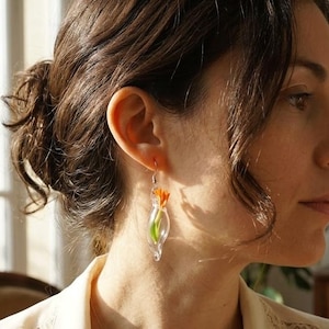 Pu&ograve; includere: Un orecchino a goccia in vetro trasparente contenente un piccolo fiore arancione brillante con uno stelo verde. L'orecchino &egrave; dotato di un gancio in metallo color argento e presenta un design delicato e trasparente a forma di ampolla.