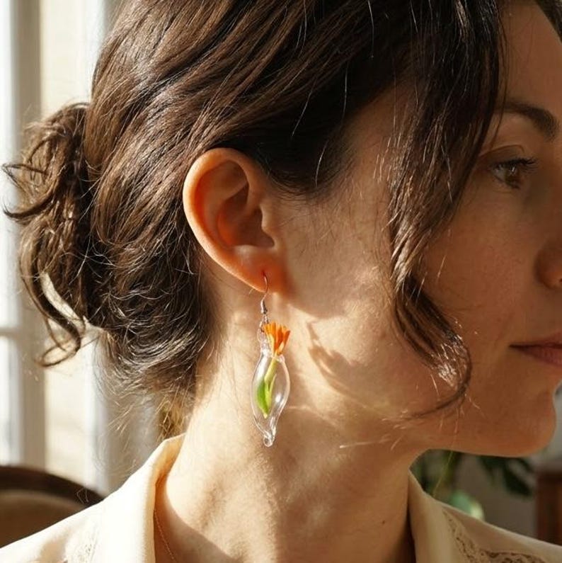 Pu&ograve; includere: Un orecchino a goccia in vetro trasparente contenente un piccolo fiore arancione brillante con uno stelo verde. L'orecchino &egrave; dotato di un gancio in metallo color argento e presenta un design delicato e trasparente a forma di ampolla.