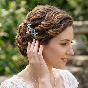 May include: A hair accessory featuring three small, light blue enamelled flowers with thin, gold-toned stems and leaves. The floral piece is secured into a wavy, brown updo hairstyle, adding a delicate touch of colour to the hair arrangement.