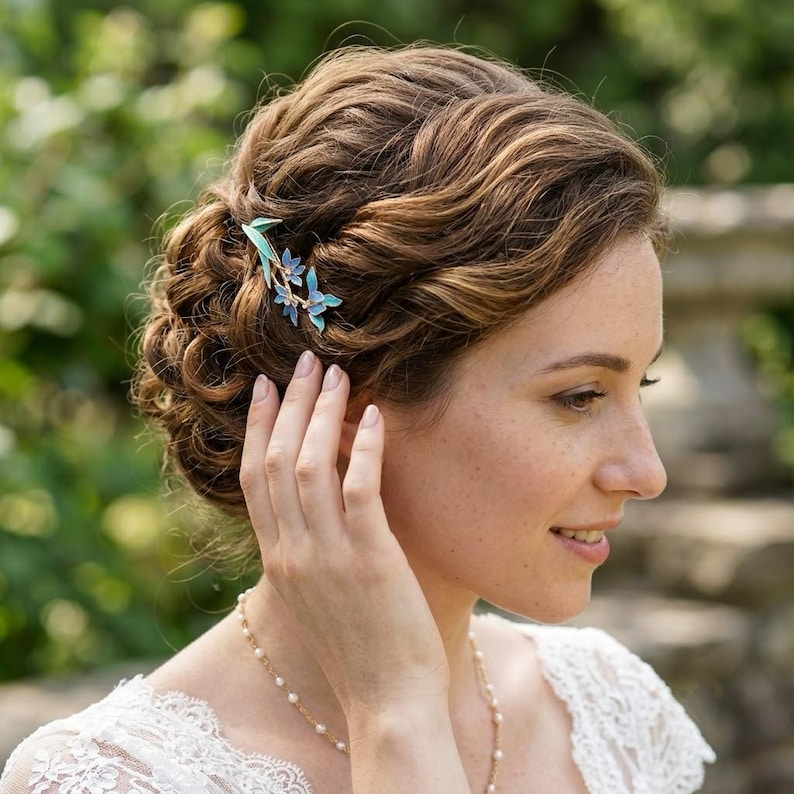 May include: A hair accessory featuring three small, light blue enamelled flowers with thin, gold-toned stems and leaves. The floral piece is secured into a wavy, brown updo hairstyle, adding a delicate touch of colour to the hair arrangement.