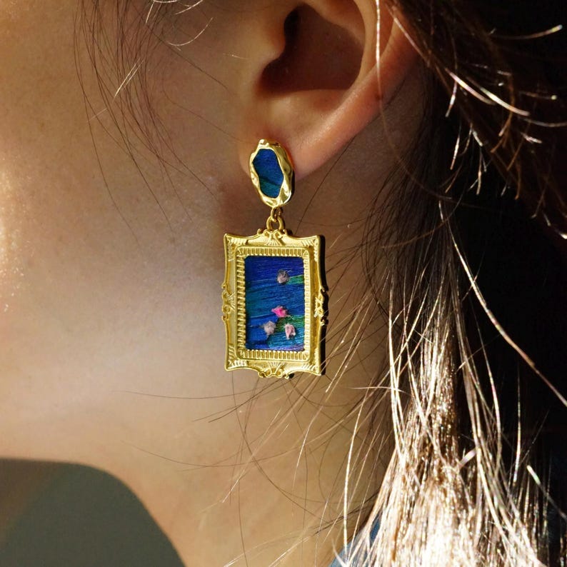 May include: A gold-toned drop earring featuring a rectangular frame with ornate detailing. The center displays a miniature painting of a blue water scene with small pink flowers. A small, irregular blue stone is set at the top of the earring.