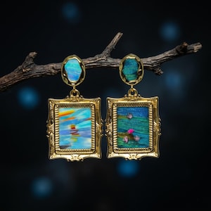 May include: A pair of gold-toned rectangular frame earrings featuring ornate edges. The left earring displays a blue and orange abstract landscape, while the right shows a green textured surface with small pink accents. Both have oval tops.