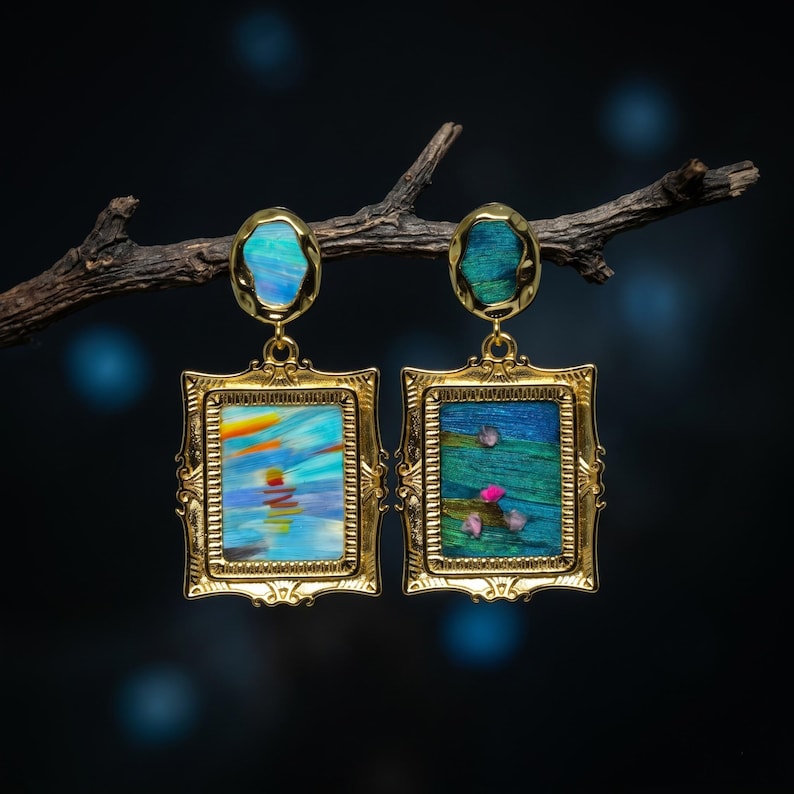 May include: A pair of gold-toned rectangular frame earrings featuring ornate edges. The left earring displays a blue and orange abstract landscape, while the right shows a green textured surface with small pink accents. Both have oval tops.