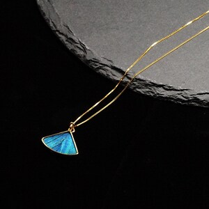 May include: A gold-toned necklace with a pendant featuring a blue, fan-shaped design. The pendant has a gold border and a delicate chain. The necklace is displayed against a dark background.
