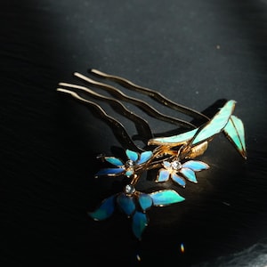 May include: A gold-toned hair comb featuring three wavy prongs and a decorative floral arrangement. The flowers have blue and teal enamel petals with small clear crystals at their centres, accompanied by elongated green enamel leaves.