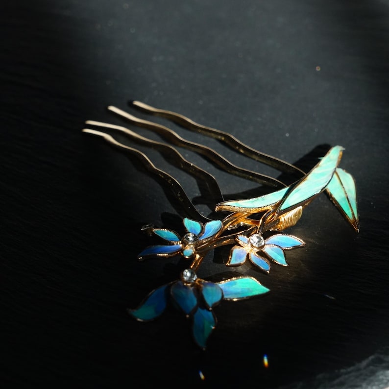 May include: A gold-toned hair comb featuring three wavy prongs and a decorative floral arrangement. The flowers have blue and teal enamel petals with small clear crystals at their centres, accompanied by elongated green enamel leaves.
