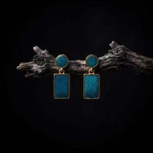 May include: A pair of drop earrings featuring a gold-toned metal frame surrounding a textured, deep teal rectangular base topped with a smaller circular teal element. The earrings are suspended from a dark, weathered wooden branch against a black background.