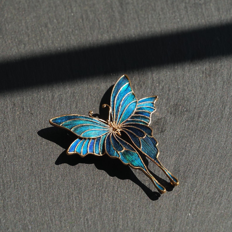 May include: A butterfly brooch with a gold-coloured frame and detailed blue and teal enamel wings. The butterfly's delicate design is highlighted by the gold outline, creating a striking contrast. A beautiful accessory.