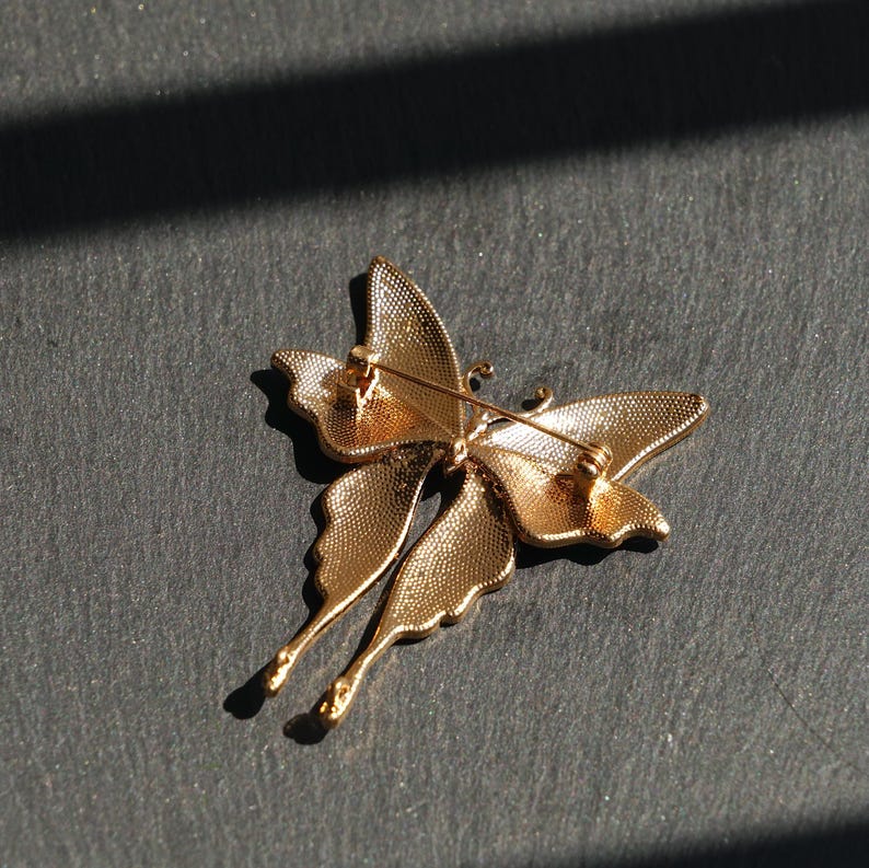 May include: A gold butterfly brooch with intricate wing detailing. The metal brooch features a pin fastening on the reverse. The butterfly's wings have a textured finish, and the design is elegant. The brooch is a decorative accessory.