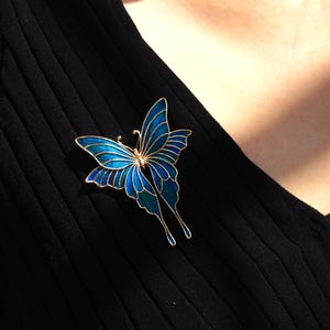 May include: A gold butterfly brooch with blue and teal enamel. The brooch is pinned to black ribbed fabric. The butterfly design features outstretched wings and delicate antennae.