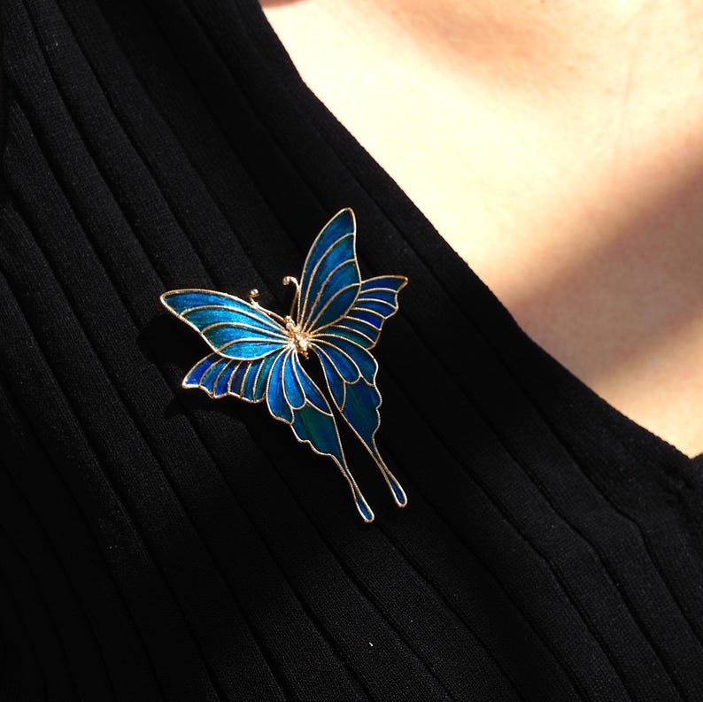 May include: A gold butterfly brooch with blue and teal enamel. The brooch is pinned to black ribbed fabric. The butterfly design features outstretched wings and delicate antennae.