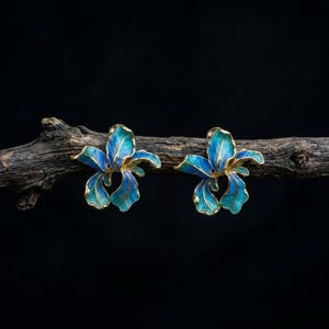 Könnte beinhalten: Ein Paar Ohrstecker in Form von Irisblüten mit leuchtend blauen und türkisfarbenen Emaille-Blütenblättern und zarten goldfarbenen Rändern. Die Ohrringe sind auf einem strukturierten, dunkelbraunen Holzzweig vor schwarzem Hintergrund platziert.