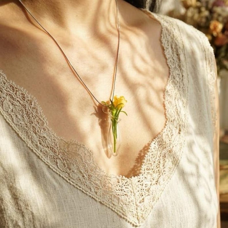 Pu&ograve; includere: Una collana delicata con una sottile catena metallica e un piccolo ciondolo a forma di fiala in vetro trasparente. All'interno del contenitore di vetro &egrave; visibile un singolo fiore giallo brillante con steli verdi, sospeso come elemento botanico.