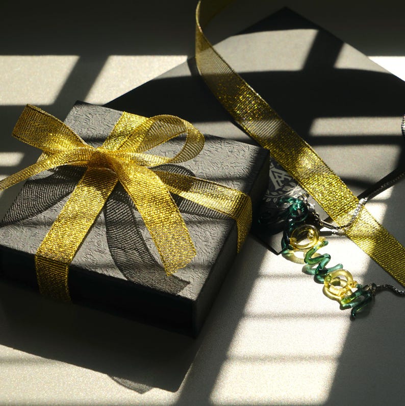Pode incluir: Uma caixa de presente preta amarrada com uma fita dourada, com uma fita combinando e um colar com detalhes verdes e dourados. A cena &eacute; iluminada pela luz do sol, criando sombras.