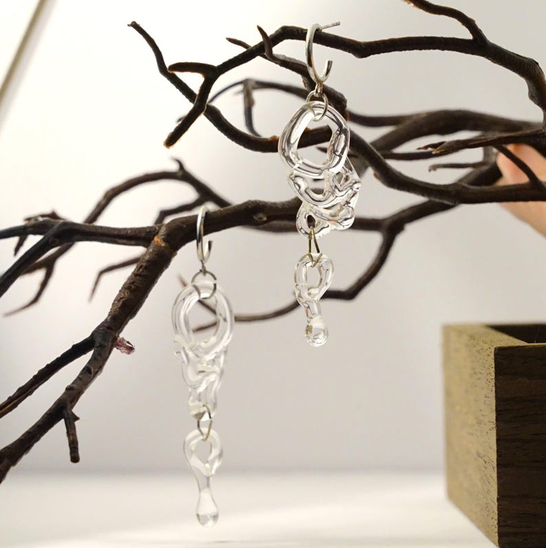 Pode incluir: Brincos pendurados transparentes com um design abstrato &uacute;nico. Os brincos s&atilde;o feitos de um material transparente, possivelmente vidro ou acr&iacute;lico, e apresentam uma s&eacute;rie de formas interconectadas. Penduram-se em ganchos prateados, criando um visual moderno e art&iacute;stico.