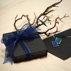 May include: A square black gift box tied with a dark blue sheer ribbon bow. Beside the box are two trapezoidal earrings with blue and green striped patterns, a gold ring featuring a small blue butterfly, and a dark, bare, branching twig.