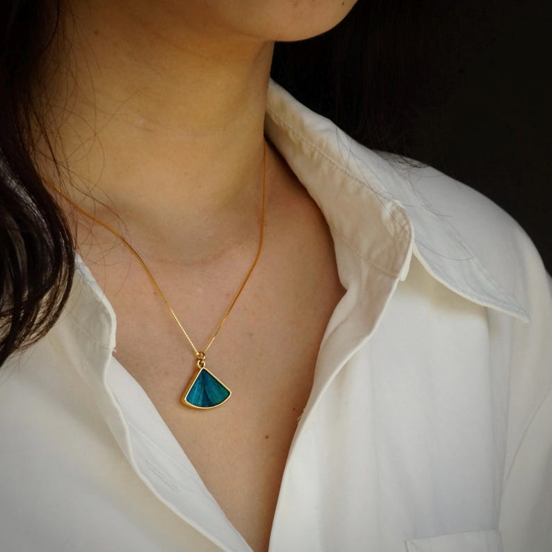 May include: A gold-colored necklace with a teal pendant. The pendant is a triangular shape with a gold border. The necklace is worn over a white collared shirt.