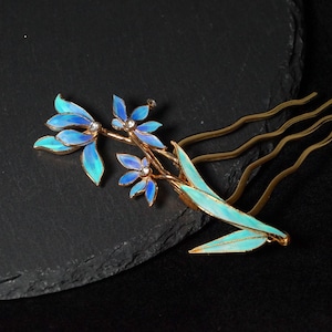 May include: A gold-toned hair comb featuring an intricate floral design with iridescent blue and turquoise enamel petals. The centre of each flower is accented with a small, clear crystal. The comb has three wavy prongs for secure placement.