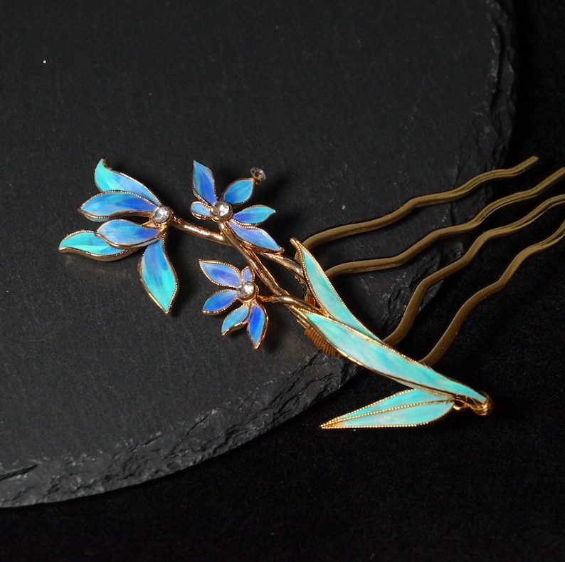 May include: A gold-toned hair comb featuring an intricate floral design with iridescent blue and turquoise enamel petals. The centre of each flower is accented with a small, clear crystal. The comb has three wavy prongs for secure placement.