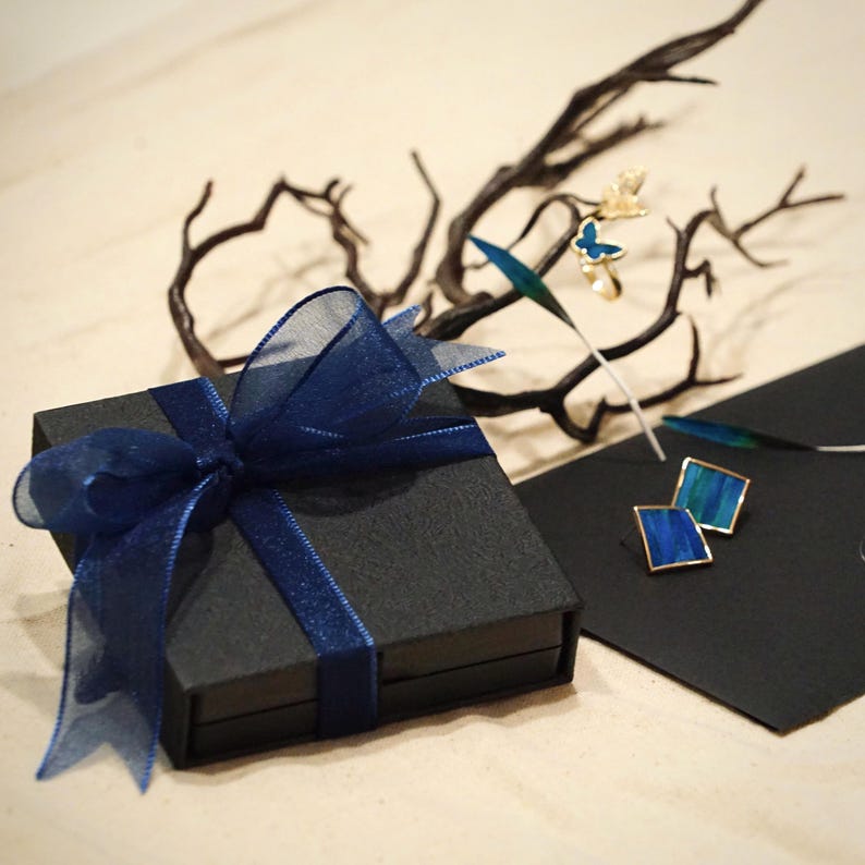 May include: A black gift box tied with a sheer navy blue ribbon. The box is next to a black card with square blue and gold earrings. A gold butterfly ring and decorative branches are in the background.