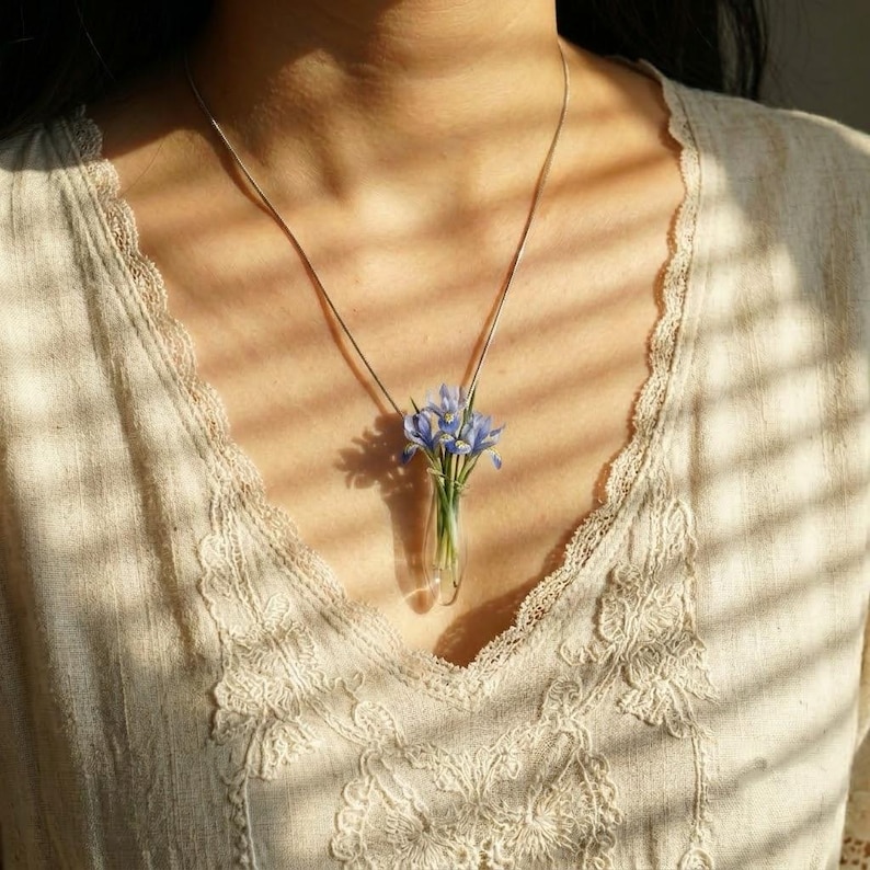 Pu&ograve; includere: Una collana con ciondolo a forma di fiala in vetro trasparente contenente un piccolo mazzo di delicati fiori di iris blu. Il ciondolo pende da una sottile catenina color argento, appoggiato su un indumento color crema con dettagli in pizzo.
