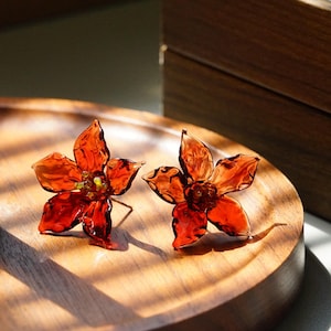 Puede incluir: Un par de pendientes con forma de flor. Los pendientes están hechos de un material translúcido, de color rojo anaranjado, con cinco pétalos. El centro de cada flor tiene un pequeño detalle verde y amarillo. Los pendientes se exhiben en una bandeja de madera.