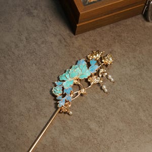Pode incluir: Um gancho de cabelo de metal dourado com um design floral intrincado com p&eacute;talas de esmalte azul e turquesa. O topo decorativo inclui pequenas contas brancas pendentes e detalhes de folhas met&aacute;licas fixadas num alfinete longo e fino.