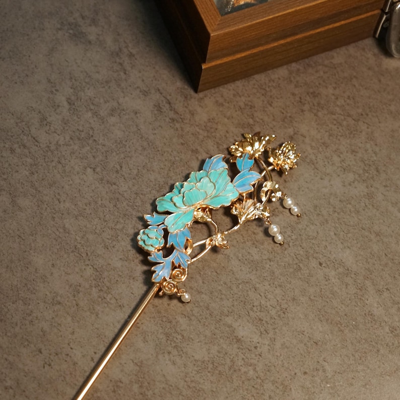 Pode incluir: Um gancho de cabelo de metal dourado com um design floral intrincado com p&eacute;talas de esmalte azul e turquesa. O topo decorativo inclui pequenas contas brancas pendentes e detalhes de folhas met&aacute;licas fixadas num alfinete longo e fino.