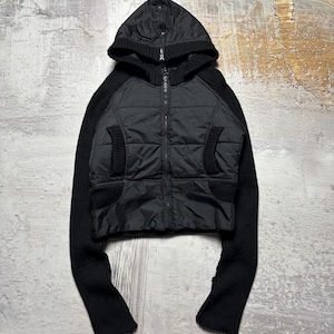 Könnte beinhalten: Schwarze Kapuzenjacke mit gesteppter Front und langen Strickärmeln. Die Jacke hat einen durchgehenden Reißverschluss und zwei Vordertaschen. Die Kapuze ist ebenfalls aus Strick. Die Jacke wird auf einer strukturierten, neutral getönten Oberfläche präsentiert.