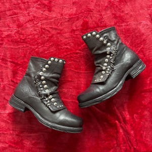 May include: A pair of black ankle boots with silver-colored stud and loop embellishments. The boots have a low heel and a slightly scrunched design on the upper shaft. The boots are displayed on a red velvet surface.