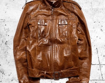 Könnte beinhalten: Eine braune Lederjacke mit Stehkragen, durchgehendem Frontreißverschluss und zwei Brusttaschen mit Druckknopfverschluss. Die Jacke verfügt über Schulterklappen mit Metallösen, einen Taillengürtel mit Schnalle und Druckknopfmanschetten.