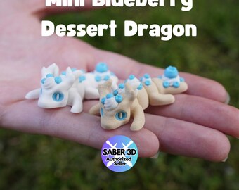 Pode incluir: Pequenas figuras de dragões em creme e bege claro com detalhes azuis. O texto "Mini Blueberry Dessert Dragon" está acima. A imagem também inclui um logotipo "SABER 3D Authorized Seller". As figuras estão deitadas.