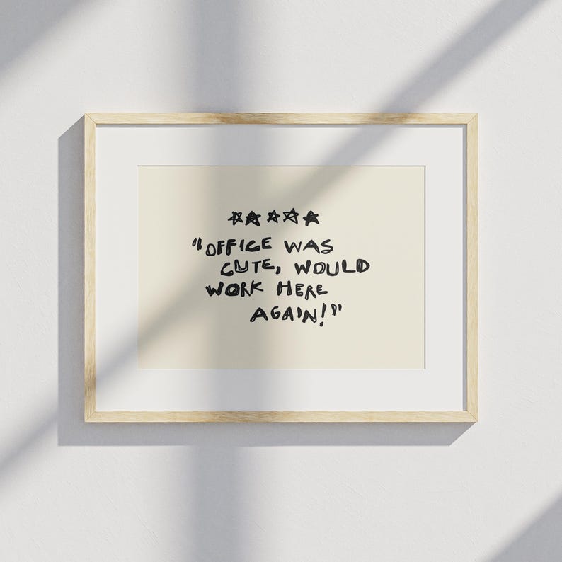 May include: A framed print with a light beige background and a wooden frame. The print features the text "OFFICE WAS CUTE, WOULD WORK HERE AGAIN!" in black, handwritten-style font, with five stars above the text.