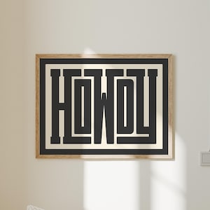 May include: A framed art print displaying the word "HOWDY" in a bold, black, stylized font. The text is set against a cream background, framed by a black border and a natural wood frame. The artwork is mounted on a white wall.