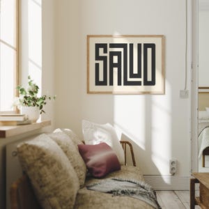 Puede incluir: Un cuadro enmarcado con la palabra "SALUD" en letras geom&eacute;tricas negras y audaces. La obra de arte se muestra sobre un fondo crema, enmarcada en madera clara. El cuadro cuelga sobre un sof&aacute; con cojines decorativos.