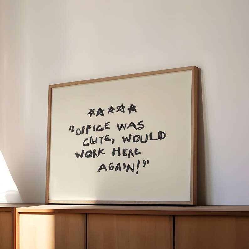 May include: A framed print with the text "Office was cute, would work here again!" in black handwritten style. The print has a cream-colored background and a light brown wooden frame. Above the text are four black stars.