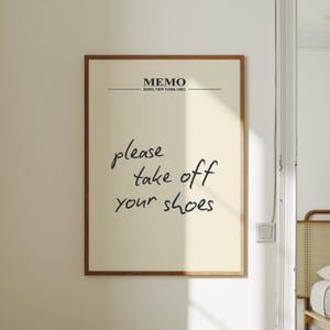 May include: A framed print with the text "please take off your shoes" in a handwritten style. The print also includes the word "MEMO" and the location "SOHO, NEW YORK 10012" in a minimalist font. The frame is a warm brown colour.