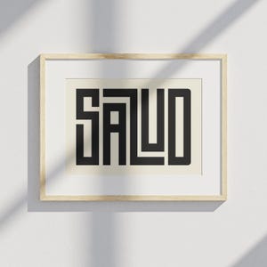 Puede incluir: Un estampado enmarcado con la palabra "SALUD" en letras geom&eacute;tricas negras y en negrita. El estampado est&aacute; sobre un fondo crema y enmarcado en un marco de madera clara. La obra de arte se exhibe en una pared blanca.