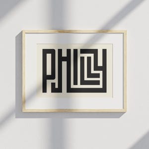Könnte beinhalten: Gerahmter Kunstdruck mit dem Wort "PHILLY" in einer kräftigen, geometrischen schwarzen Schrift auf cremefarbenem Hintergrund. Das Kunstwerk ist in einem hellen Holzrahmen eingefasst, geeignet für die Wanddekoration. Der Druck wird an einer weißen Wand präsentiert.