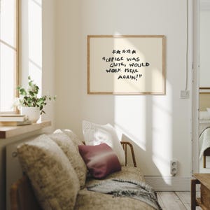 May include: A framed print with the text "Office was cute, would work here again!" in black lettering. The print hangs above a sofa with patterned cushions and a burgundy pillow. A window with sunlight and a plant is visible.