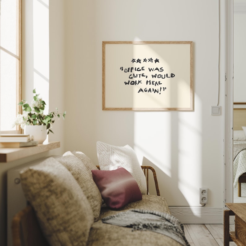 May include: A framed print with the text "Office was cute, would work here again!" in black lettering. The print hangs above a sofa with patterned cushions and a burgundy pillow. A window with sunlight and a plant is visible.