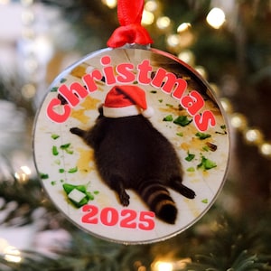 May include: Circular Christmas bauble featuring a raccoon in a Santa hat. The red text reads "Christmas" and "2025". The bauble is suspended by a red ribbon.