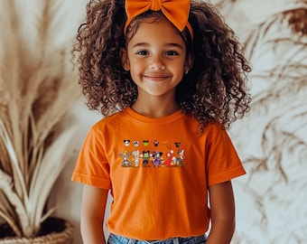 May include: An orange short-sleeved t-shirt featuring a graphic print of various cartoon characters dressed in Halloween costumes, paired with a matching orange fabric bow headband. The shirt displays a row of small character icons above the main figures.
