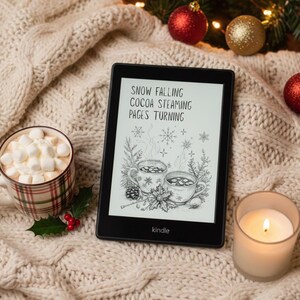 May include: A Kindle e-reader displaying the text "Snow Falling Cocoa Steaming Pages Turning" with a drawing of two mugs of cocoa. A mug of cocoa with marshmallows, a lit candle, and Christmas ornaments rest on a cable-knit blanket.