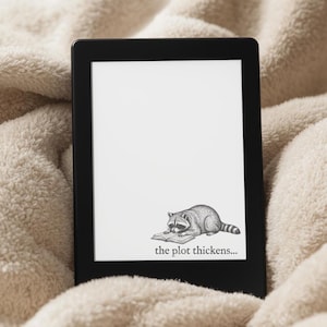 May include: A black e-reader displays a grayscale illustration of a raccoon reading a book, with the text "the plot thickens...". The e-reader is set against a soft, cream-colored blanket, suggesting a cozy reading environment.
