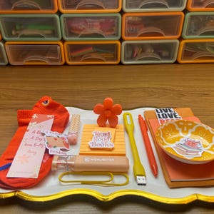 Op de afbeelding: Een verzameling kantoorartikelen op een wit dienblad met gouden rand, waaronder een oranje sok, een bladwijzer met tekst, een bloemvormige clip, een notitieblok met een Read Good Books-sticker, een markeerstift, een USB-lampje en een klein schaaltje.