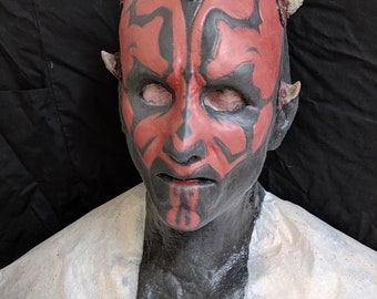 Op de afbeelding: Een gedetailleerd masker van een personage met een rood en zwart gezichtsontwerp, met hoorns en een strenge uitdrukking. Het masker staat tegen een donkere achtergrond en toont de ingewikkelde details van de kenmerken van het personage.
