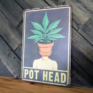 May include: A rectangular metal sign with a dark blue background features a person with a terracotta pot for a head, with a cannabis plant growing out of it. The sign reads "POT HEAD" in yellow letters.